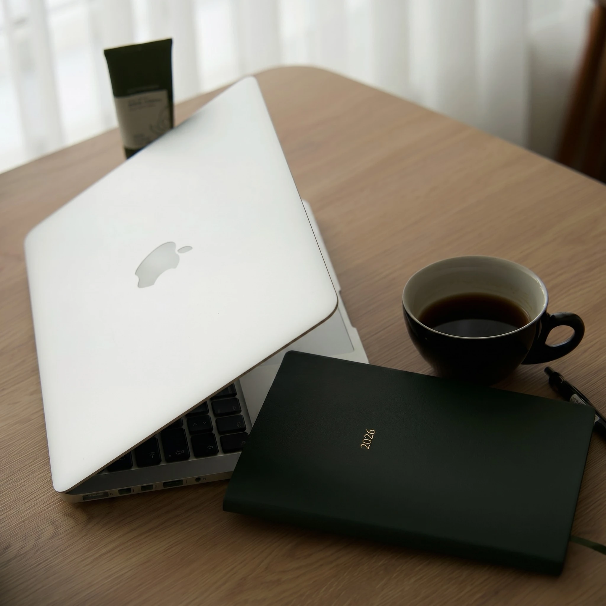 Laptop, coffee, and a 2026 notebook on a warm wooden table beside soft curtains.